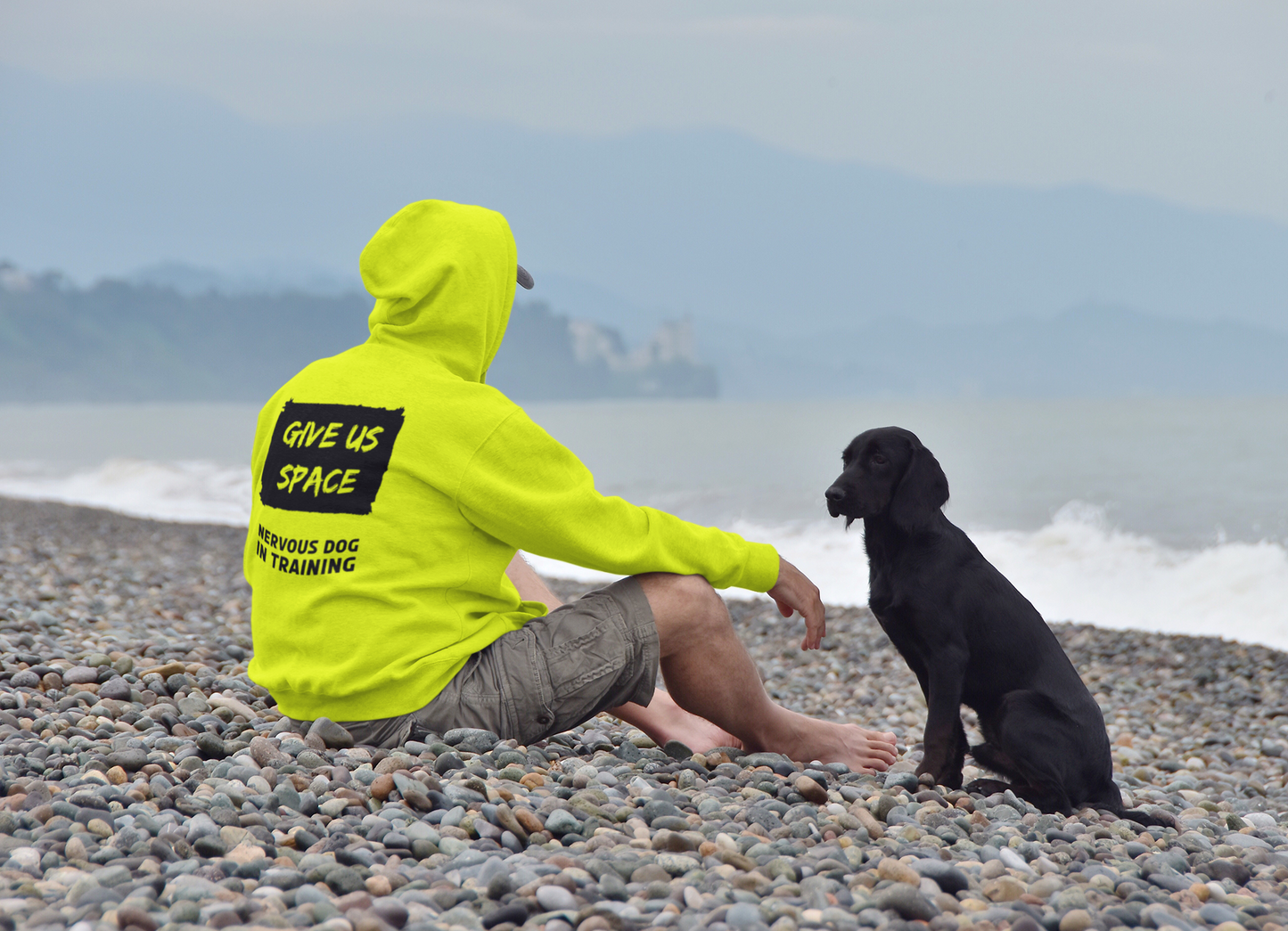 man-sitting-at-a-beach-in-a-yellow--give-us-space-hoody-with-his-nervous-black-dog